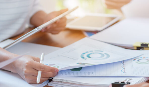Close-up of hands reviewing printed data charts with a tablet in the background.