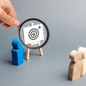 Magnifying glass focuses on a bullseye target graphic on an easel beside wooden figures.