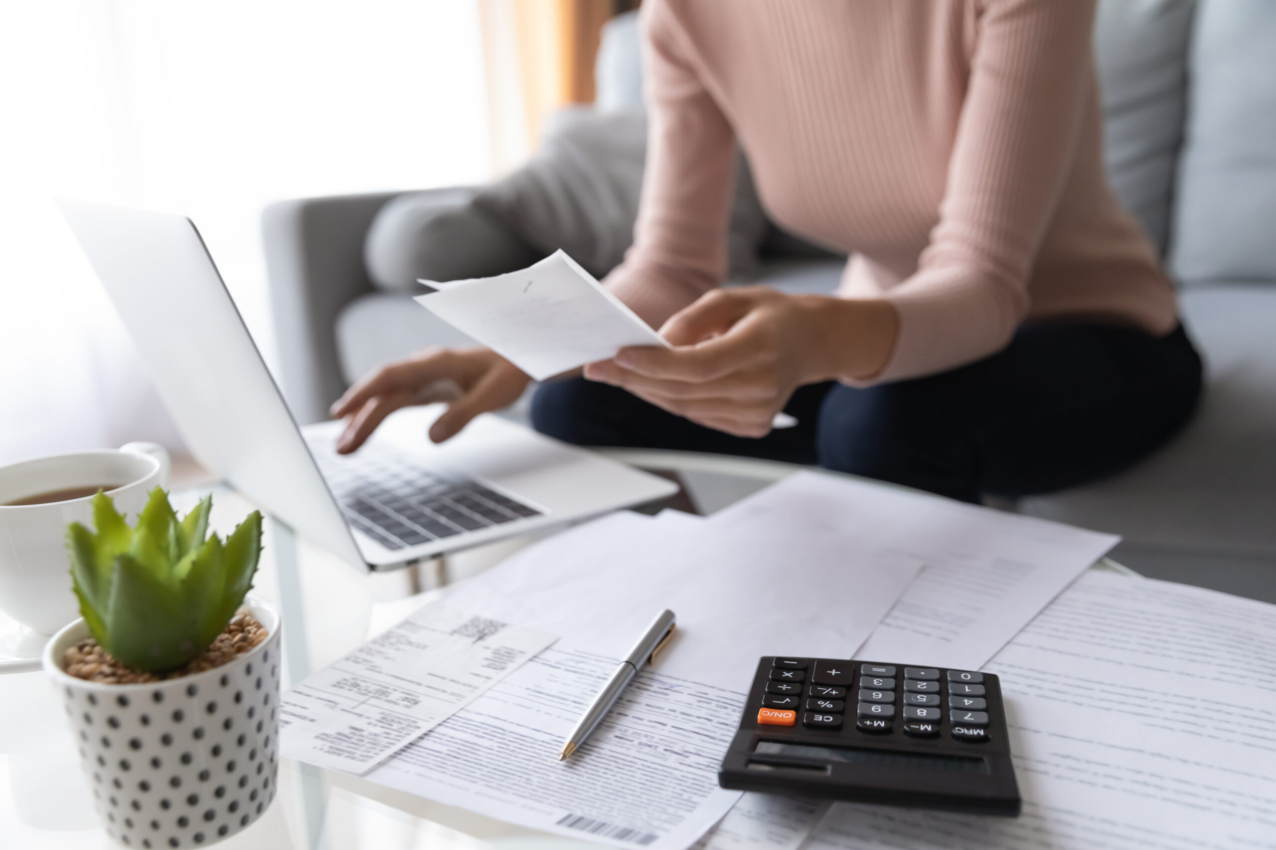 Woman manages household finances on a laptop, receipts and calculator spread across a coffee-table workspace.