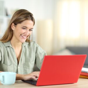 Smiling young woman works on a red laptop at home with a coffee mug nearby.