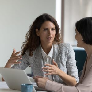 Two professional women talk animatedly around a laptop during an office meeting.