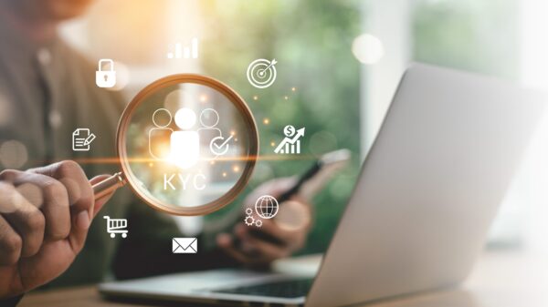 Hand holding a magnifying glass over a laptop with glowing KYC and fintech icons.