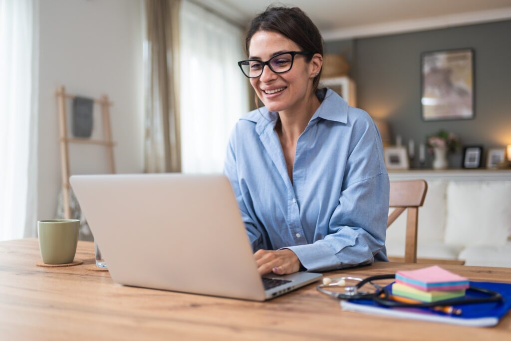 Smiling woman in glasses working on a laptop at a home table with a coffee mug.