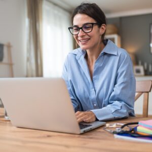 Smiling woman in glasses working on a laptop at a home table with a coffee mug.