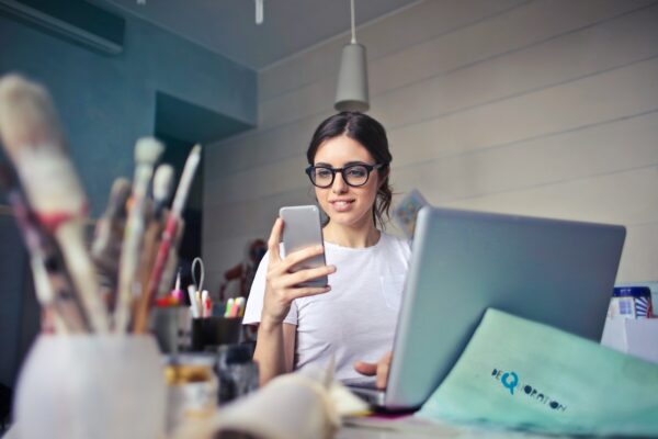 Young woman in glasses checks her phone at a creative desk cluttered with art supplies and an open laptop.