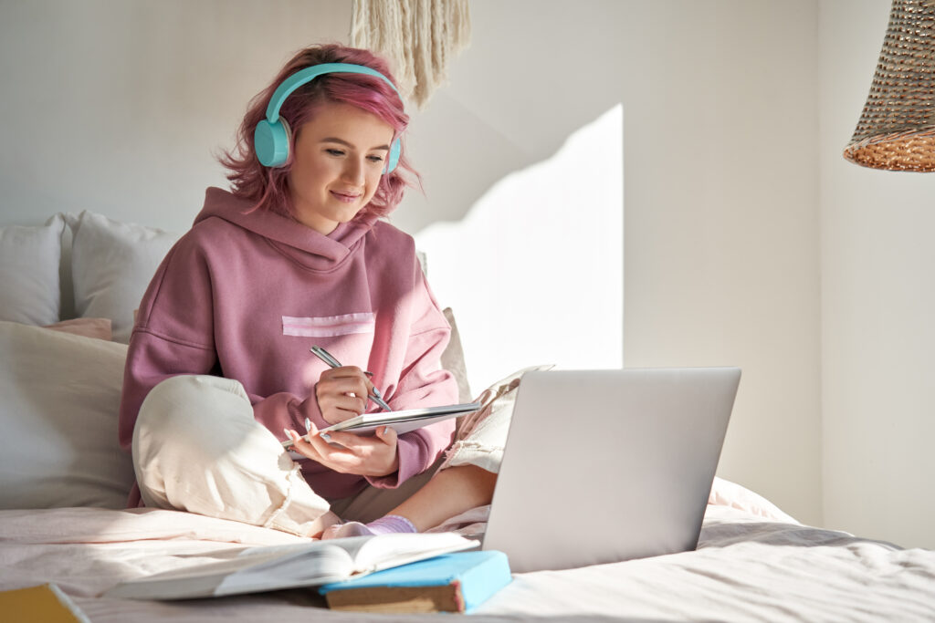 Teen with pink hair and turquoise headphones studies on a bed with a laptop and notebook.