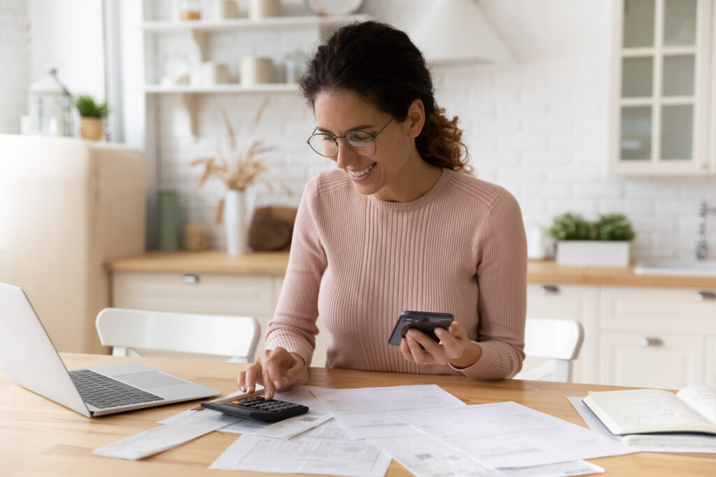 Smiling woman in glasses calculates bills with phone, calculator, and laptop at a kitchen table.