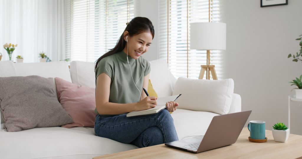 Woman on a sofa taking notes in a notebook while watching her laptop, earbuds in.