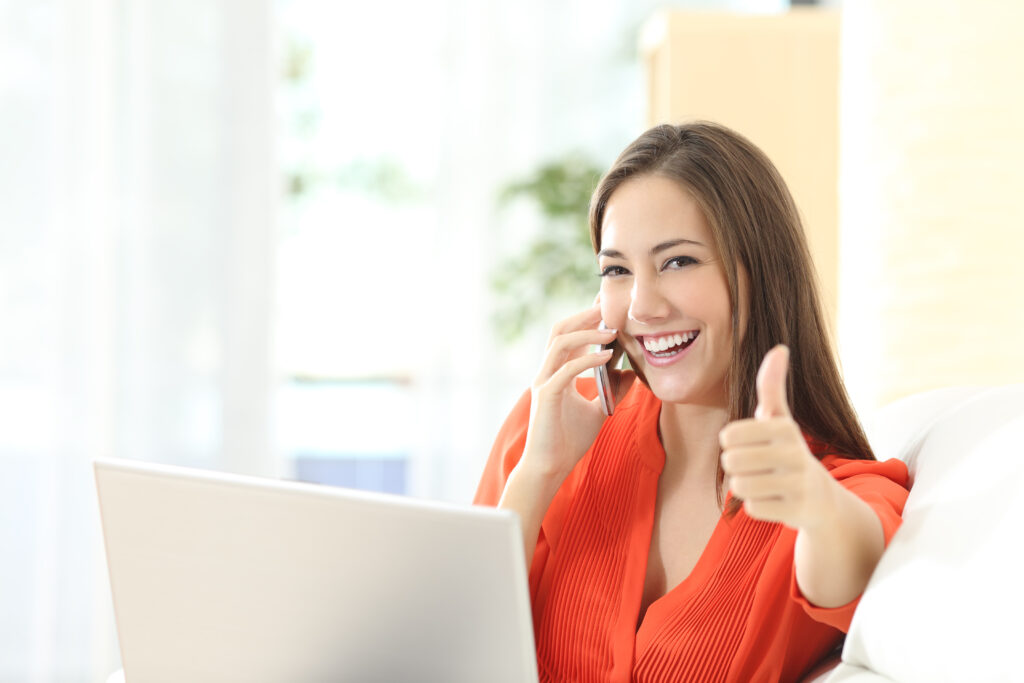 Happy woman in an orange top giving a thumbs-up while talking on the phone beside a laptop.