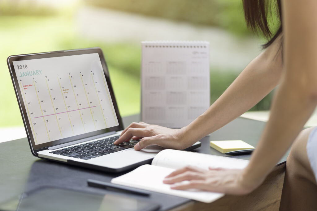 Hands updating a laptop calendar next to a planner and sticky notes on a desk.