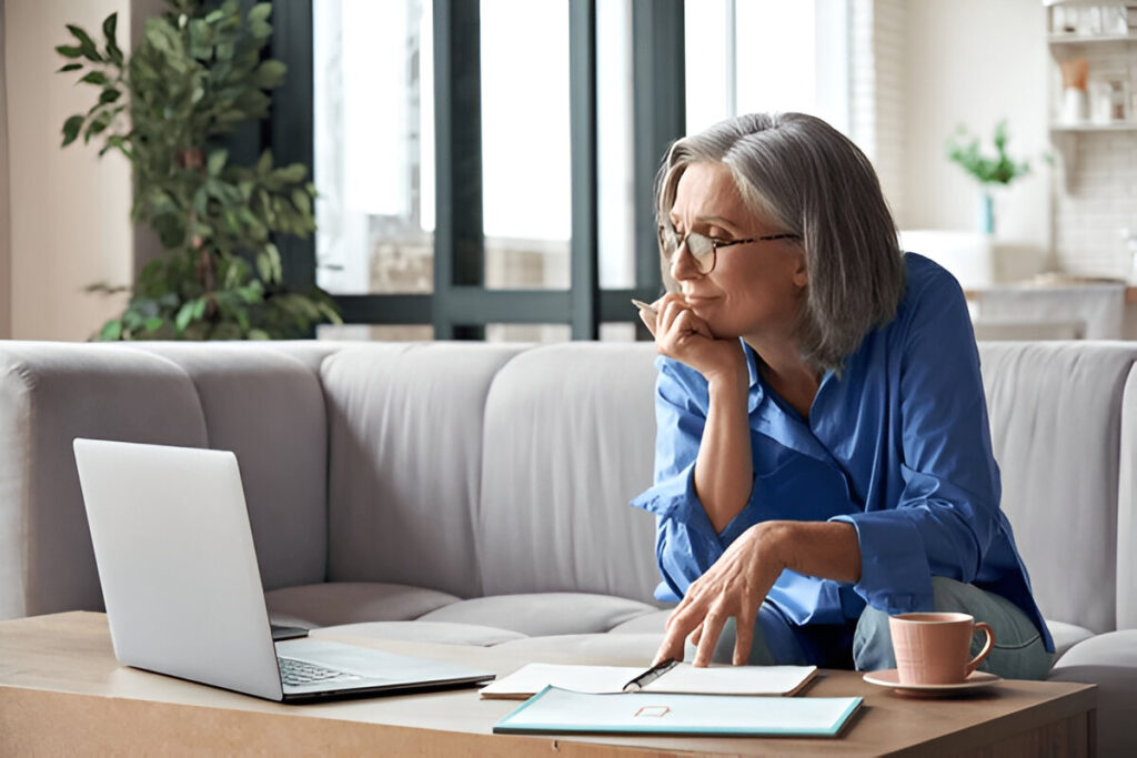 Mature woman with gray hair reviewing notes beside a laptop on a living-room coffee table.