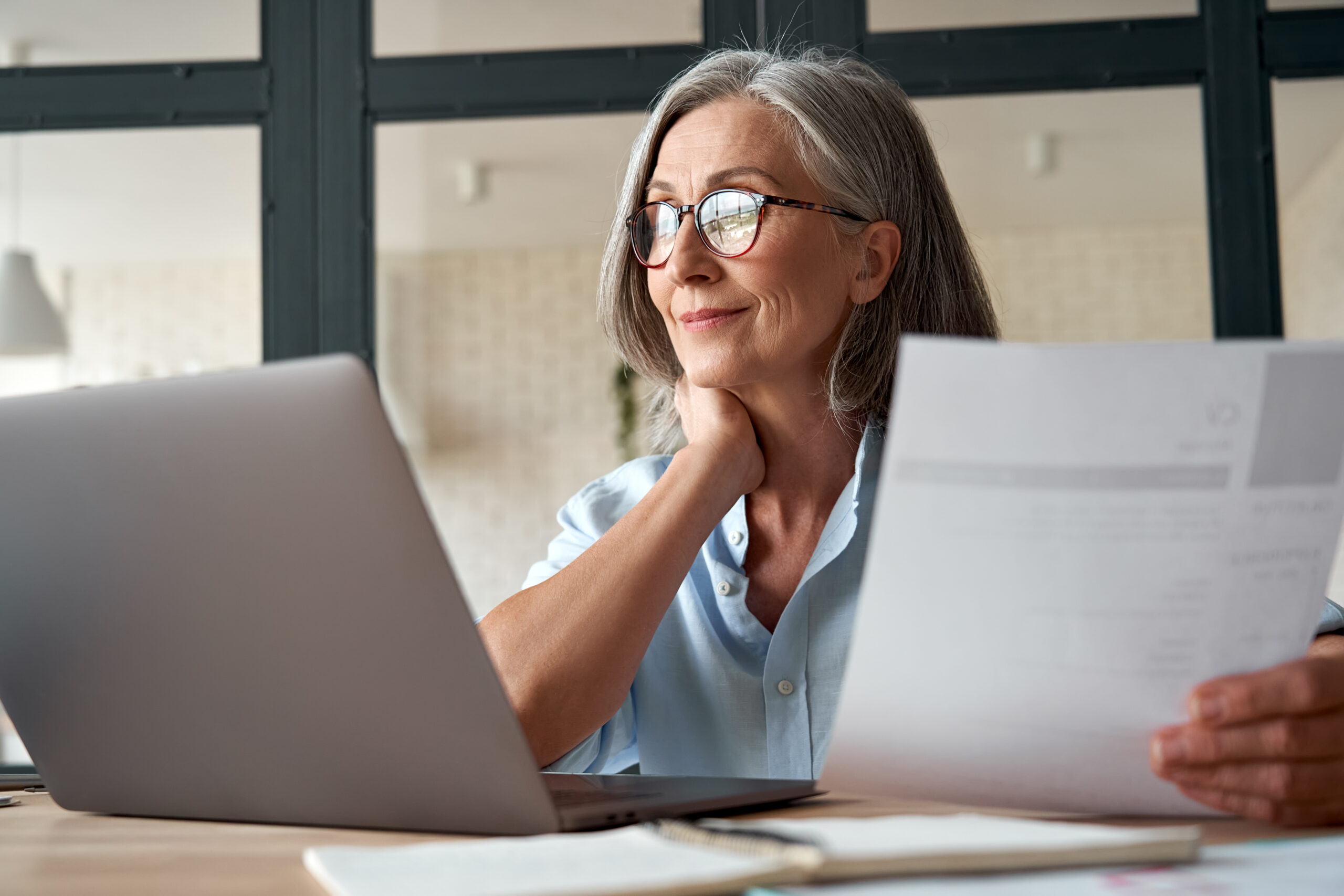 Gray-haired woman reviews paperwork while working on her laptop, resting her chin on one hand.
