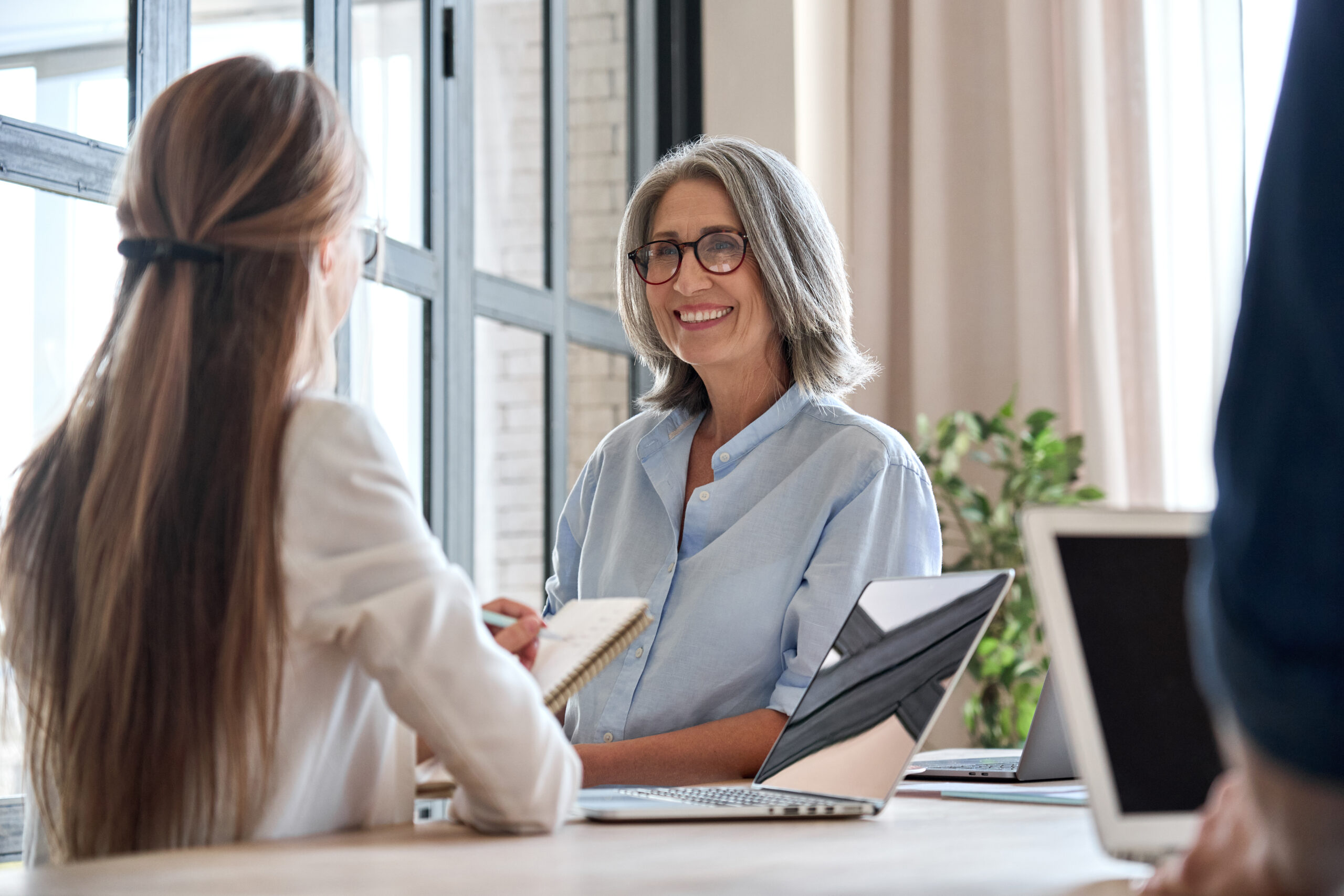 Senior professional in glasses smiles during a meeting with colleagues and open laptops.