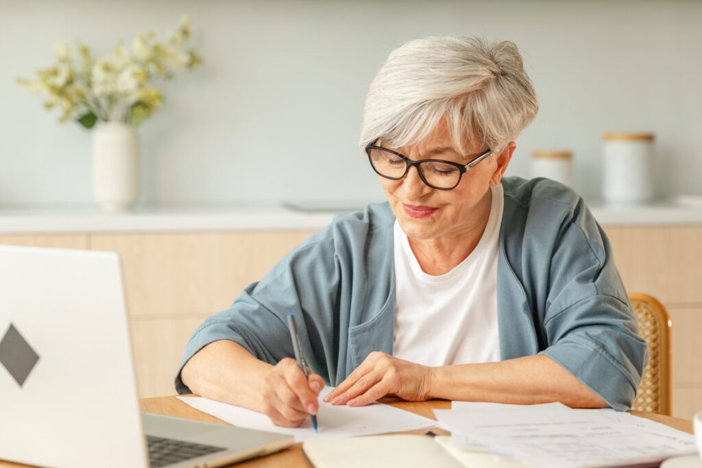 Senior woman with a silver pixie cut writes notes next to a laptop and scattered documents.