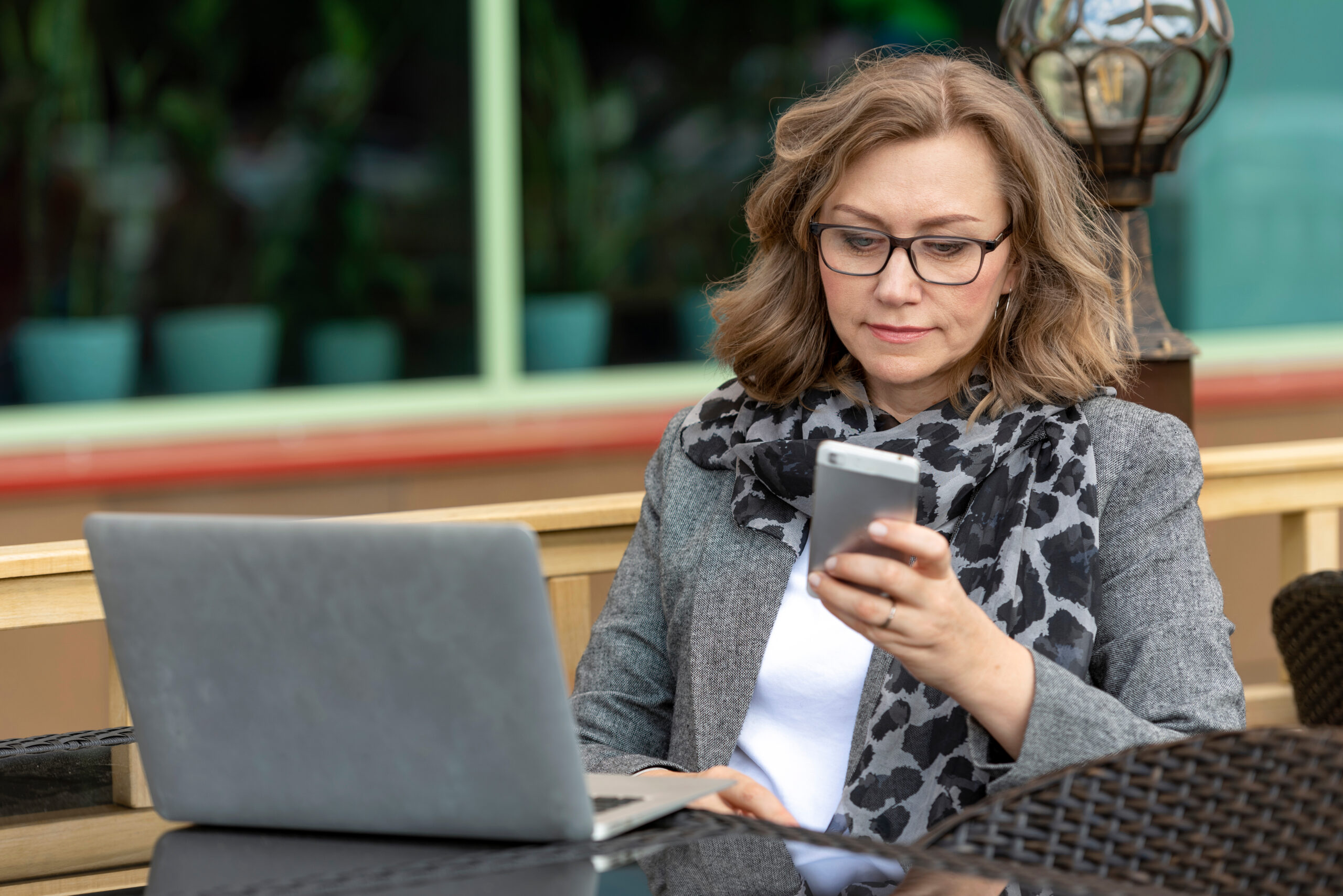 Woman in glasses at an outdoor café table checks her phone beside an open laptop.