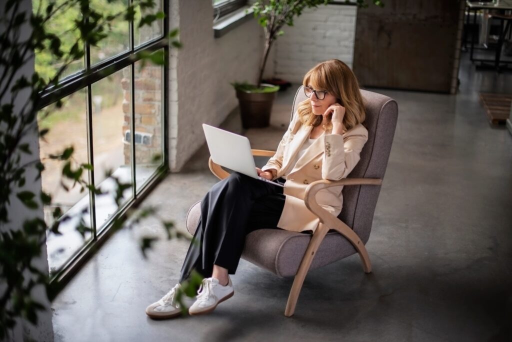 Woman seated in an armchair by tall windows concentrates on her laptop.