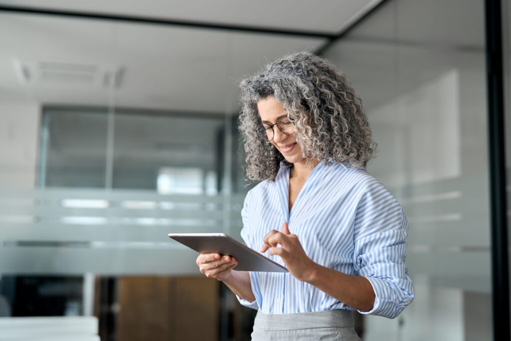 Gray-curly-haired professional reviews content on a tablet inside a glass-walled office.